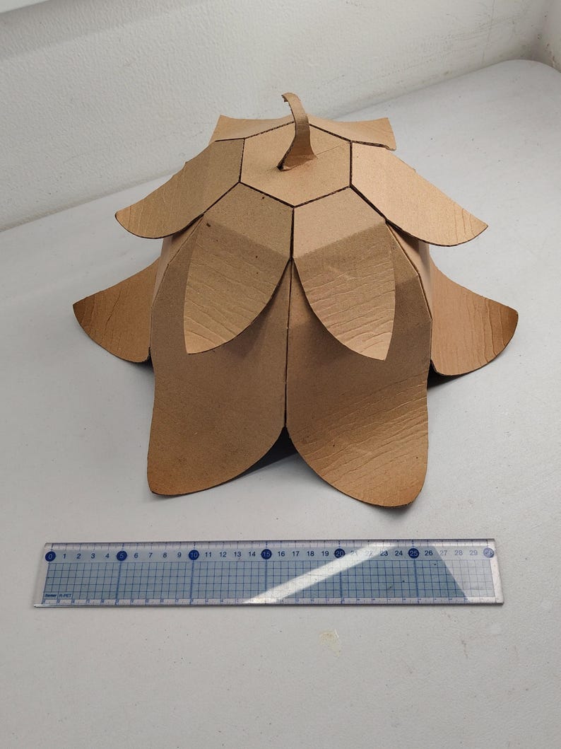 May include: A brown cardboard sculpture shaped like a flower with layered petals and a central stem, positioned behind a clear plastic ruler that measures approximately 11.5 inches in length, showing the object's scale on a white surface.