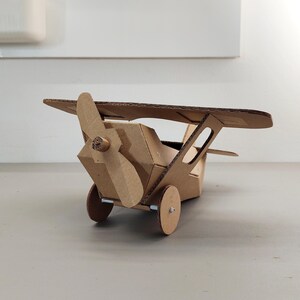 May include: A model aeroplane constructed from brown corrugated cardboard. The aircraft features a single propeller at the front, a main wing positioned above the fuselage, and two circular wheels attached to the base with small metal axles.