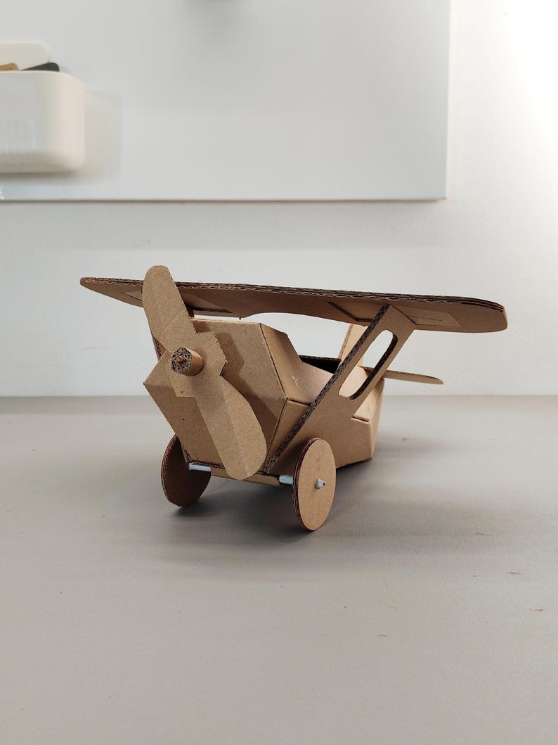 May include: A model aeroplane constructed from brown corrugated cardboard. The aircraft features a single propeller at the front, a main wing positioned above the fuselage, and two circular wheels attached to the base with small metal axles.
