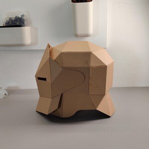 May include: A geometric, cardboard sculpture of a stylized animal head, possibly a bear or wolf, with angular planes and a dark eye slit. The sculpture is a light brown color and sits on a gray surface.