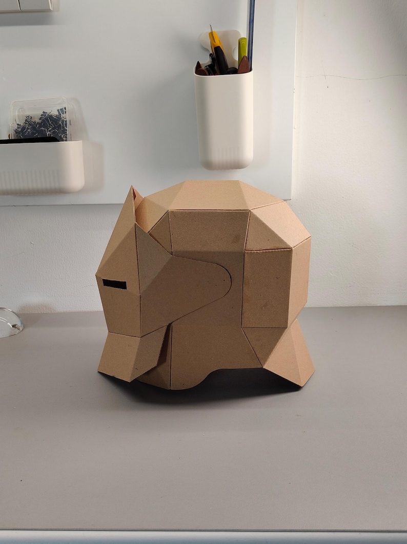 May include: A geometric, cardboard sculpture of a stylized animal head, possibly a bear or wolf, with angular planes and a dark eye slit. The sculpture is a light brown color and sits on a gray surface.