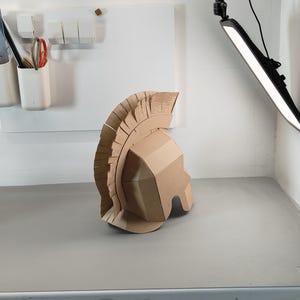 May include: A tan cardboard helmet featuring a tall, fringed crest that runs from the front to the back. The helmet has a geometric, angular design with a face guard and a prominent, layered plume made of individual vertical cardboard strips.