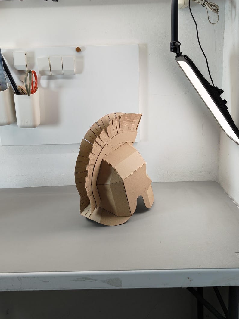 May include: A tan cardboard helmet featuring a tall, fringed crest that runs from the front to the back. The helmet has a geometric, angular design with a face guard and a prominent, layered plume made of individual vertical cardboard strips.