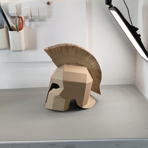 May include: A tan cardboard Spartan-style helmet featuring a prominent crest along the top. The helmet is constructed from multiple angular panels of corrugated cardboard, showcasing a distinct geometric design with a wide eye opening.