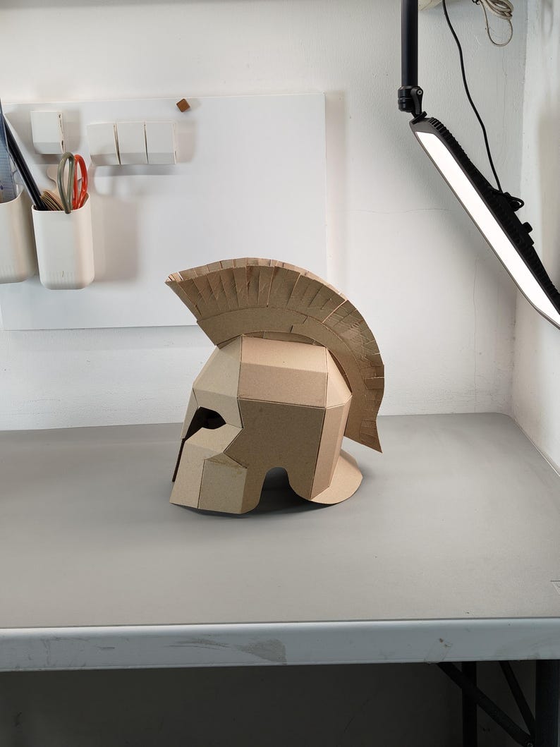 May include: A tan cardboard Spartan-style helmet featuring a prominent crest along the top. The helmet is constructed from multiple angular panels of corrugated cardboard, showcasing a distinct geometric design with a wide eye opening.
