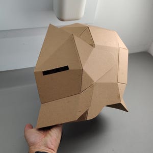May include: A geometric cardboard helmet with a brown finish. The helmet features a series of angular planes and a horizontal eye slit. The design suggests a futuristic or abstract aesthetic, suitable for cosplay or artistic projects.