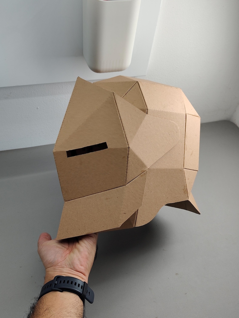May include: A geometric cardboard helmet with a brown finish. The helmet features a series of angular planes and a horizontal eye slit. The design suggests a futuristic or abstract aesthetic, suitable for cosplay or artistic projects.