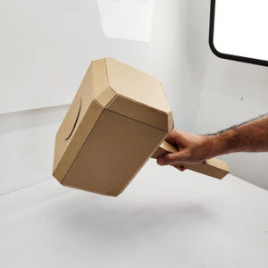 May include: A cardboard model of Thor's hammer, Mjolnir, with a brown finish. The hammer is held by a hand, with the handle extending to the right.