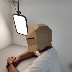 May include: A person wearing a geometric cardboard mask with a rectangular eye slit. The mask is tan and covers the entire head. The person is wearing a white t-shirt and a black watch. The background is a white wall and a gray table.