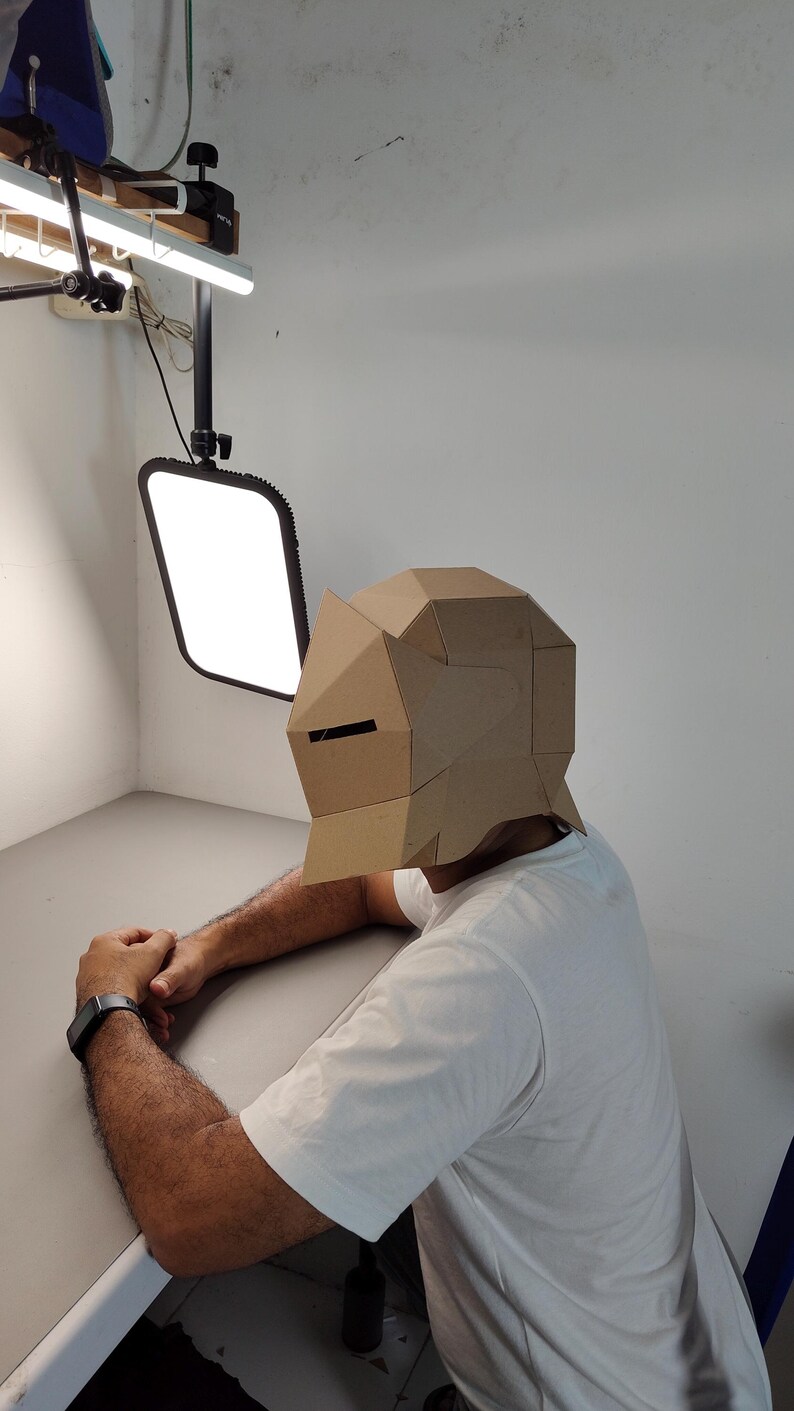 May include: A person wearing a geometric cardboard mask with a rectangular eye slit. The mask is tan and covers the entire head. The person is wearing a white t-shirt and a black watch. The background is a white wall and a gray table.