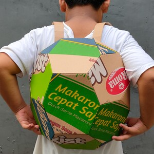 K&ouml;nnte beinhalten: Ein Kind, das ein wei&szlig;es Hemd tr&auml;gt, tr&auml;gt einen Rucksack aus Pappkartons. Der Rucksack ist mit den Worten "Makaroni Cepat Saji" und "Quick Serve Macaroni" in Gr&uuml;n und Gelb verziert. Der Rucksack ist auch mit einem rot-wei&szlig;en Logo verziert, auf dem "Roma" steht.