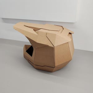 May include: A geometric helmet constructed from brown cardboard, featuring a faceted, angular design with a prominent visor and a large open face area. The structure is composed of multiple interlocking panels, creating a rigid, multi-sided shape.