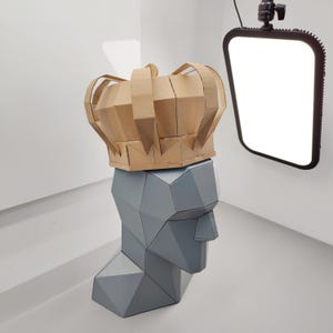 Puede incluir: Una escultura de cabeza geométrica gris con una corona de papel marrón. La corona está hecha de cartón plegado y se asienta sobre la cabeza. La escultura está en una habitación blanca con una luz brillante.
