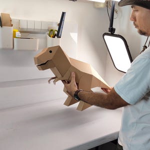 May include: A geometric T-Rex dinosaur model constructed from light brown cardboard. The figure features a faceted, angular design with a distinct head, small arms, a long tail, and two legs. The surface is plain, matte, and tan in colour.
