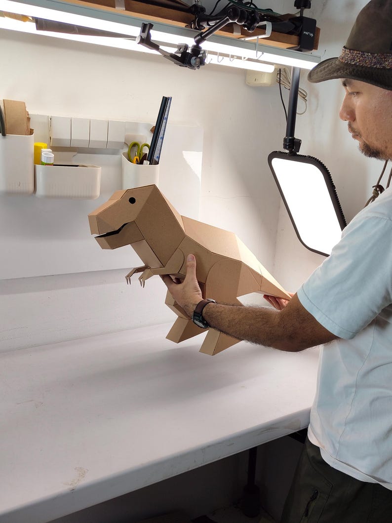 May include: A geometric T-Rex dinosaur model constructed from light brown cardboard. The figure features a faceted, angular design with a distinct head, small arms, a long tail, and two legs. The surface is plain, matte, and tan in colour.