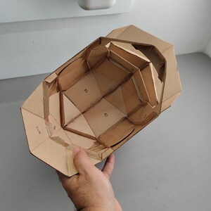 May include: A partially constructed cardboard model with geometric shapes and visible seams. The model is held by a hand, showcasing its interior structure and the numbered sections. The cardboard is a natural brown color.