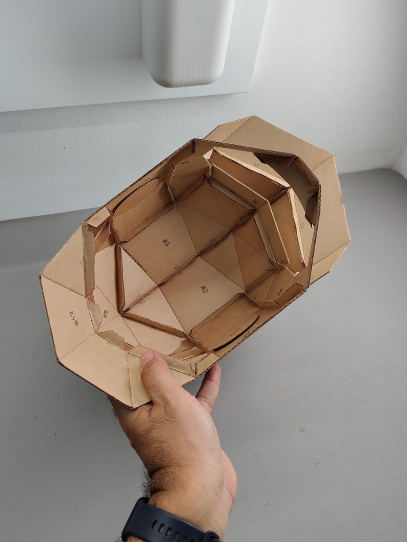 May include: A partially constructed cardboard model with geometric shapes and visible seams. The model is held by a hand, showcasing its interior structure and the numbered sections. The cardboard is a natural brown color.