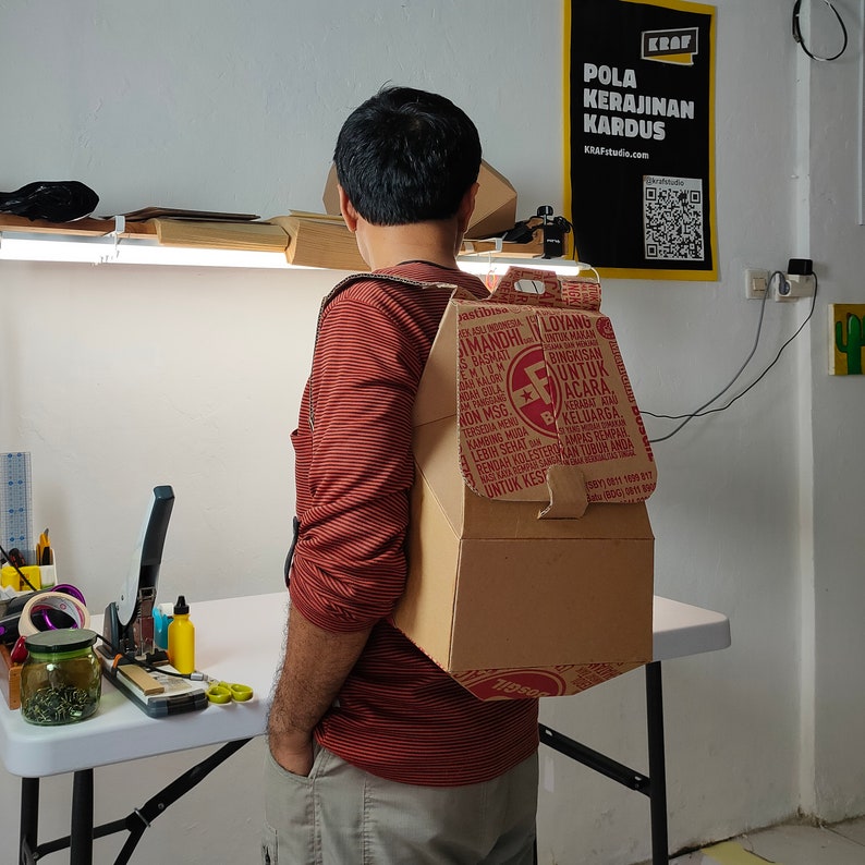 May include: A person wearing a red and white striped shirt is wearing a cardboard box backpack. The box is decorated with red and white text and a red star logo. The text on the box reads "IMANDHI LOYANG SBASMATT PREMIUM AN PINGGANG WON MSG VINTUK MAKAN BINGKISAN UNTUK ACARA KERABAT KONG CAMPING MEKELUARGA MUDA LEBIH SEHAT UNTUK KESE AMPAS REMPAR KAN TUBURA 119se".