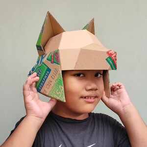 May include: A child wearing a handmade cardboard helmet. The helmet is constructed from recycled cardboard boxes, featuring geometric shapes and angular designs. The cardboard has visible text and graphics from the original packaging, with a natural brown color and green accents.