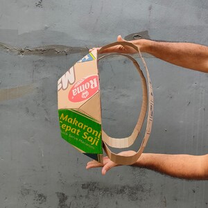 K&ouml;nnte beinhalten: Ein Kartonring, hergestellt aus einer Makkaroni-Schachtel. Die Schachtel tr&auml;gt die Aufschrift "Makaroni Cepat Saji" und "Quick Serve Macaroni". Der Ring wird von zwei H&auml;nden gehalten.