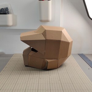 Op de afbeelding: Een geometrische helm gemaakt van bruin karton, met een gefacetteerd, hoekig ontwerp met een opvallend viziergedeelte en een afgeronde achterkant. De structuur bestaat uit meerdere platte, onderling verbonden panelen in een helmvorm.