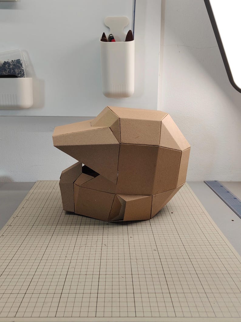Op de afbeelding: Een geometrische helm gemaakt van bruin karton, met een gefacetteerd, hoekig ontwerp met een opvallend viziergedeelte en een afgeronde achterkant. De structuur bestaat uit meerdere platte, onderling verbonden panelen in een helmvorm.