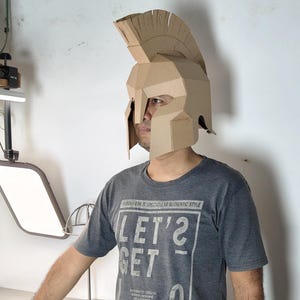 May include: A tan cardboard helmet featuring a tall crest and a face guard with eye openings. The helmet is constructed from folded panels and is worn over the head. The wearer is dressed in a dark gray t-shirt with white text that reads LET'S GET LOST.
