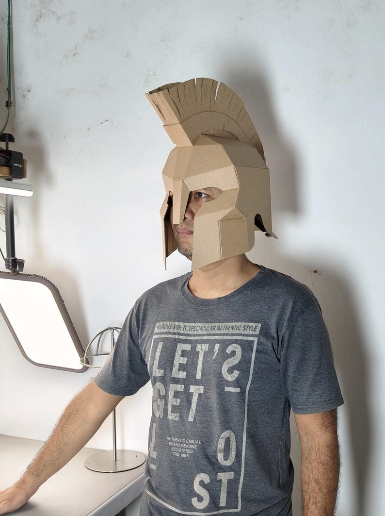 May include: A tan cardboard helmet featuring a tall crest and a face guard with eye openings. The helmet is constructed from folded panels and is worn over the head. The wearer is dressed in a dark gray t-shirt with white text that reads LET'S GET LOST.