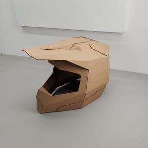 Puede incluir: Un casco de motocross de cartón marrón con visera. El casco está hecho de cartón plegado, resaltando los bordes y ángulos. Se encuentra sobre una superficie gris.