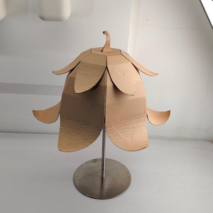May include: A table lamp featuring a shade constructed from brown corrugated cardboard, shaped like a flower with layered petals. The shade is supported by a thin, vertical silver metal rod attached to a circular, flat silver metal base.