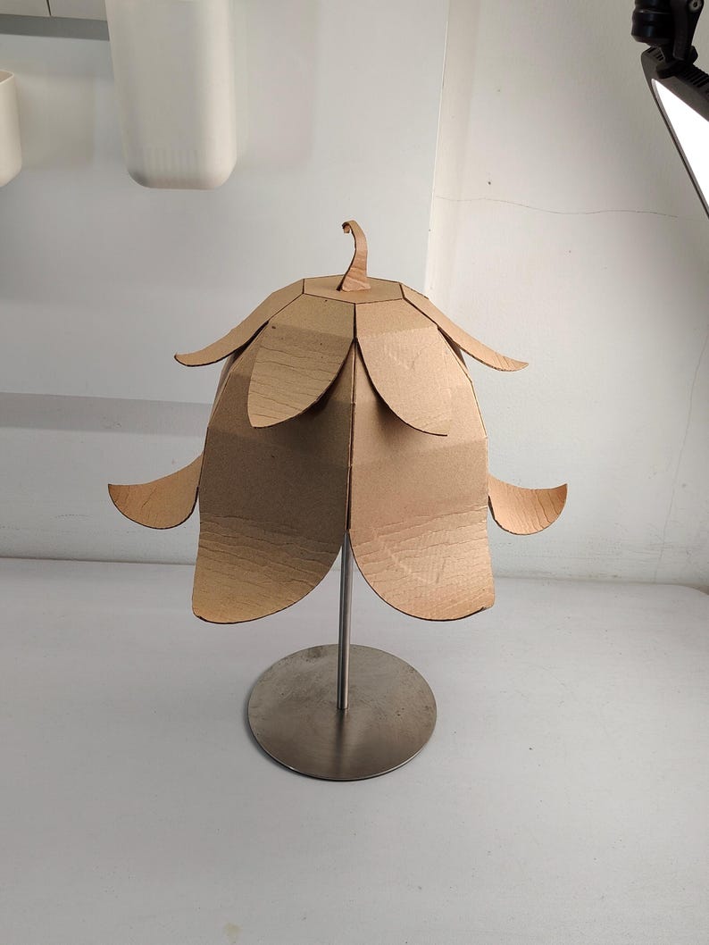 May include: A table lamp featuring a shade constructed from brown corrugated cardboard, shaped like a flower with layered petals. The shade is supported by a thin, vertical silver metal rod attached to a circular, flat silver metal base.