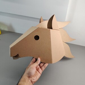 Puede incluir: Una m&aacute;scara de cabeza de caballo de cart&oacute;n color canela construida con paneles geom&eacute;tricos. La m&aacute;scara presenta una oreja puntiaguda, un recorte circular para el ojo y una crin estilizada que se extiende hacia atr&aacute;s, hecha de cart&oacute;n marr&oacute;n.