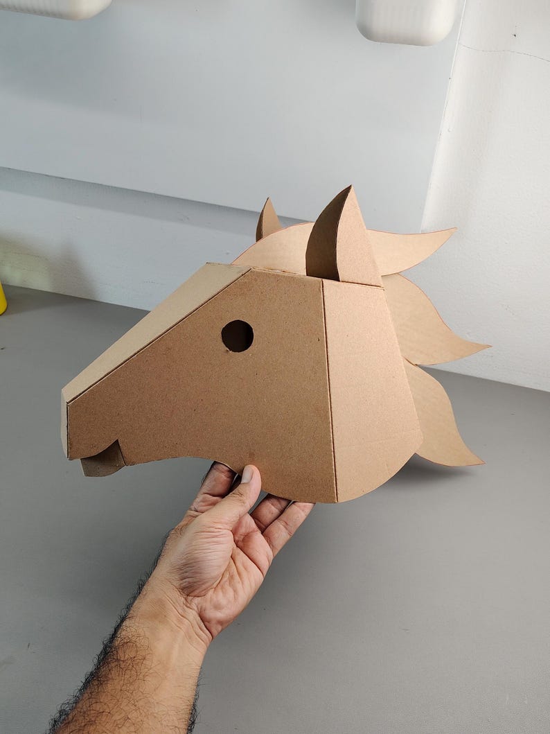 Puede incluir: Una m&aacute;scara de cabeza de caballo de cart&oacute;n color canela construida con paneles geom&eacute;tricos. La m&aacute;scara presenta una oreja puntiaguda, un recorte circular para el ojo y una crin estilizada que se extiende hacia atr&aacute;s, hecha de cart&oacute;n marr&oacute;n.