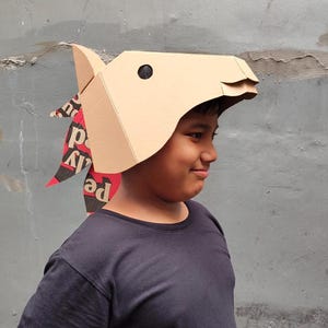 Puede incluir: Una m&aacute;scara de caballo de cart&oacute;n color canela con dise&ntilde;o geom&eacute;trico, que presenta un ojo circular negro y una crin hecha de papel reciclado con texto impreso en rojo, negro y blanco. La m&aacute;scara tiene pliegues angulares y una oreja puntiaguda.