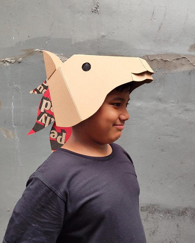 Puede incluir: Una m&aacute;scara de caballo de cart&oacute;n color canela con dise&ntilde;o geom&eacute;trico, que presenta un ojo circular negro y una crin hecha de papel reciclado con texto impreso en rojo, negro y blanco. La m&aacute;scara tiene pliegues angulares y una oreja puntiaguda.
