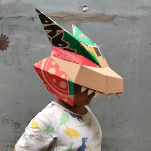 May include: A handcrafted dragon mask made from recycled cardboard. The mask is primarily brown, with red, green, and black details from pizza boxes and other packaging. The dragon's head has pointed ears and jagged teeth, creating a playful, imaginative design.