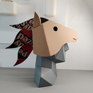 Puede incluir: Una escultura geom&eacute;trica de cabeza de caballo con hocico y orejas de cart&oacute;n color canela, combinada con una secci&oacute;n de cuello gris facetada. La crin est&aacute; hecha de material con estampado rojo y negro con el texto "ized" y "Pe" visible.