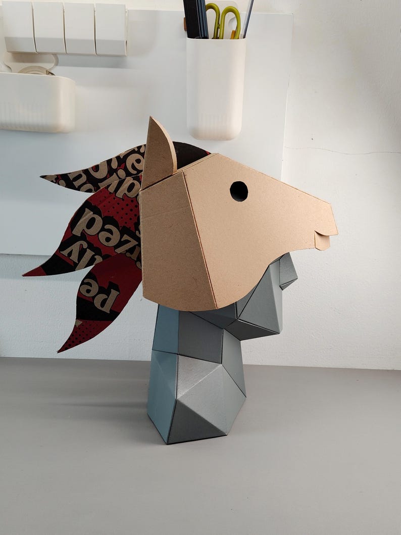 Puede incluir: Una escultura geom&eacute;trica de cabeza de caballo con hocico y orejas de cart&oacute;n color canela, combinada con una secci&oacute;n de cuello gris facetada. La crin est&aacute; hecha de material con estampado rojo y negro con el texto "ized" y "Pe" visible.