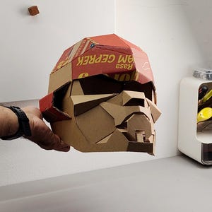May include: A geometric cardboard sculpture of a head, featuring a red and brown color scheme. The head is constructed from various pieces of cardboard, with visible text on the surface. The design appears to be a stylized representation of a face.
