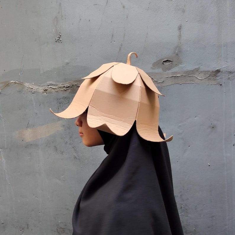 May include: A tan cardboard hat shaped like a flower with overlapping petals and a small stem at the top. The hat is worn over a black head covering that drapes down, covering the neck and shoulders, creating a contrast between the light material.