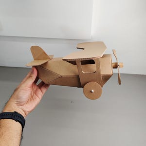 May include: A brown cardboard model aeroplane featuring a biplane design with two stacked wings, a front-mounted propeller, and circular wheels on the sides. The structure is constructed from corrugated paper material with visible edges.