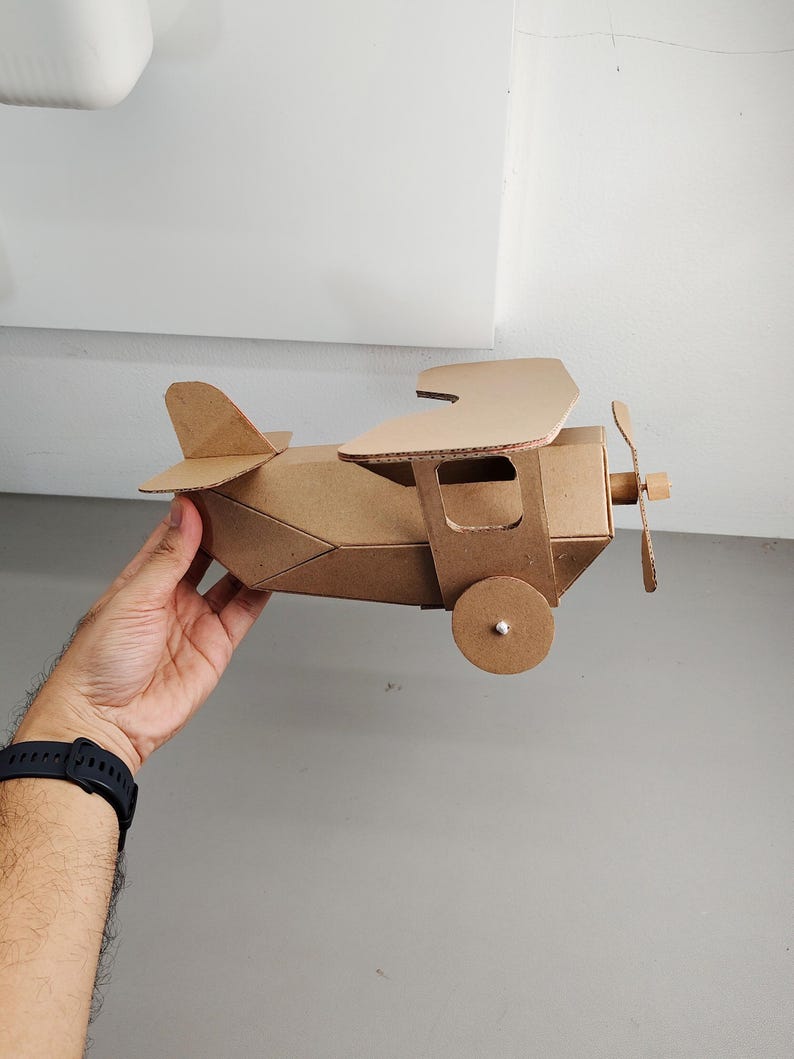 May include: A brown cardboard model aeroplane featuring a biplane design with two stacked wings, a front-mounted propeller, and circular wheels on the sides. The structure is constructed from corrugated paper material with visible edges.