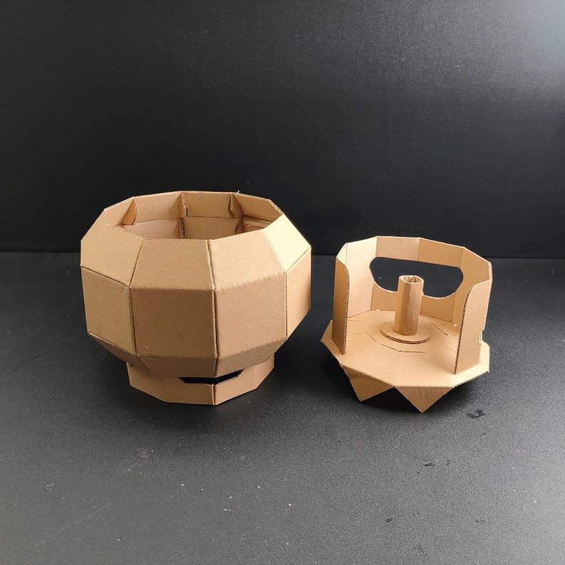 May include: A light brown cardboard container with a faceted, multi-sided exterior and a matching separate lid. The lid features a central vertical post and a handle cutout, while the main body has a wide, open top and a sturdy base structure.