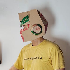 May include: A helmet constructed from brown cardboard packaging featuring green and red printed branding with the text "Mie" visible on the side. The helmet has a geometric shape with a circular cutout on the side and a yellow t-shirt underneath.
