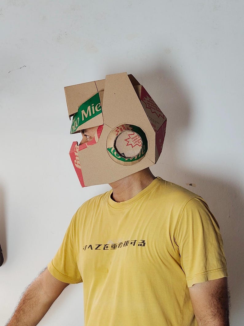 May include: A helmet constructed from brown cardboard packaging featuring green and red printed branding with the text "Mie" visible on the side. The helmet has a geometric shape with a circular cutout on the side and a yellow t-shirt underneath.