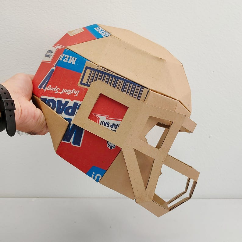 以下が含まれることがあります： シリアルボックスで作られた段ボール製のフットボールヘルメット。箱は赤、青、白で、「インスタントスパッド」と「ミール」の文字が印刷されています。