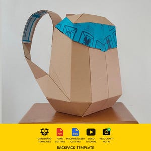 Puede incluir: Una mochila geométrica construida con cartón marrón, con una solapa superior acentuada con cinta azul impresa con iconos negros. La estructura incluye una correa para el hombro visible y un cuerpo principal con paneles angulares plegados.