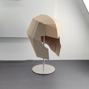 May include: A geometric helmet constructed from light brown cardboard panels, featuring a faceted, angular design. The helmet is mounted on a silver-coloured metal stand with a circular base, showcasing a structured, multi-piece assembly.