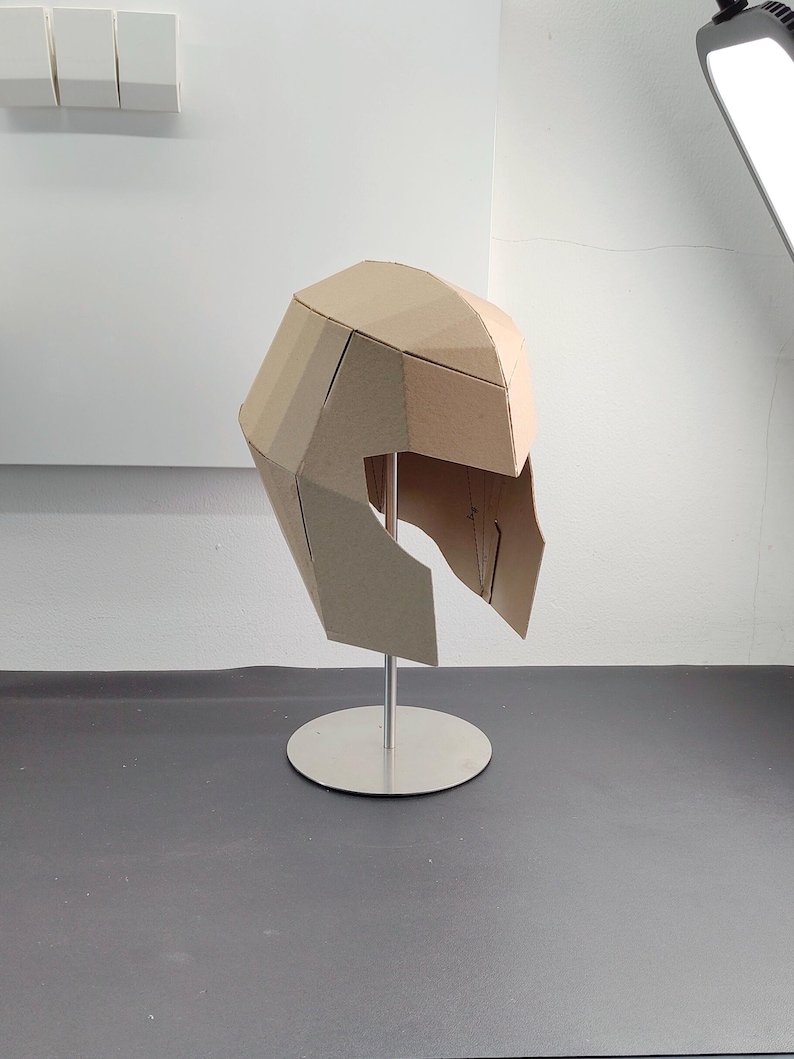 May include: A geometric helmet constructed from light brown cardboard panels, featuring a faceted, angular design. The helmet is mounted on a silver-coloured metal stand with a circular base, showcasing a structured, multi-piece assembly.