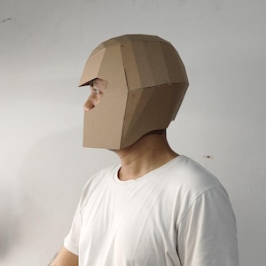May include: A geometric helmet constructed from brown cardboard panels. The helmet features a faceted, angular design with a wide visor opening and a full-coverage shell that extends down to the jawline, showcasing a clean, modular construction.